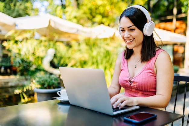 Uśmiechnięta kobieta freelancer hipster korzystająca z laptopa na tarasie restauracji podczas słuchania muzyki