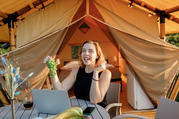 Uśmiechnięta kobieta freelancer je winogrona podczas pracy na laptopie w przytulnym namiocie glampingowym w słoneczny dzień Luksusowy namiot kempingowy na letnie wakacje na świeżym powietrzu i wakacje Koncepcja stylu życia