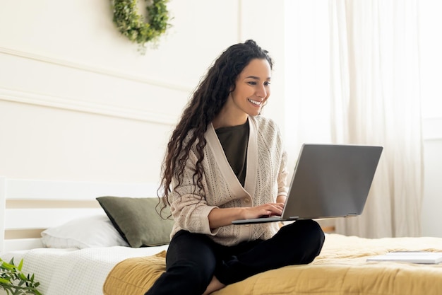 Uśmiechnięta kobieta korzystająca z laptopa leżącego na łóżku w domu