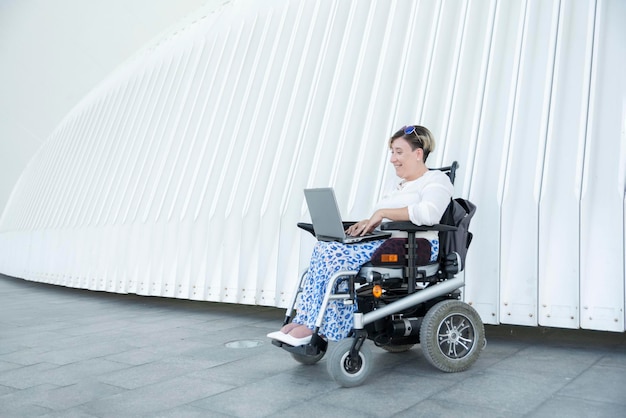 Uśmiechnięta kobieta na wózku inwalidzkim używa laptopa na zewnątrz do pracy zdalnej, zapewniając możliwości pracy osobom niepełnosprawnym