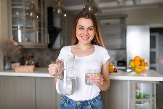Uśmiechnięta kobieta nalewa wodę pitną z dzbanka do szklanki w kuchni