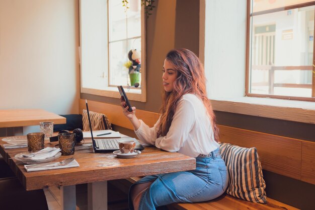 Uśmiechnięta kobieta patrząca na telefon podczas pracy w restauracji z laptopem