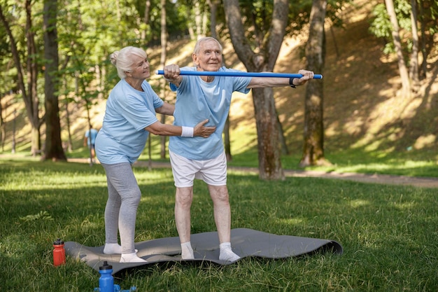Uśmiechnięta kobieta pomaga starszemu pacjentowi ćwiczyć z barem w malowniczym parku