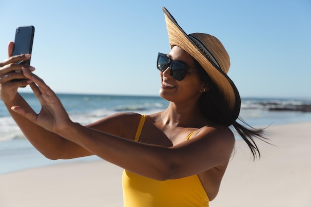 Uśmiechnięta kobieta rasy mieszanej na wakacjach na plaży, biorąc selfie. czas wypoczynku na świeżym powietrzu nad morzem.