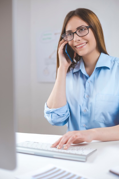 Uśmiechnięta kobieta rozmawia przez telefon, siedząc przy komputerze.