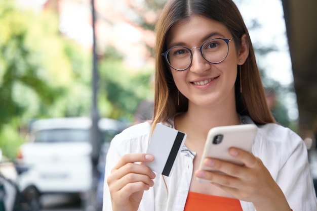 Uśmiechnięta Kobieta Trzymająca Kartę Kredytową, Korzystająca Z Telefonu Komórkowego, Robiąca Zakupy Online, Siedząca W Przytulnej Kawiarni Na świeżym Powietrzu