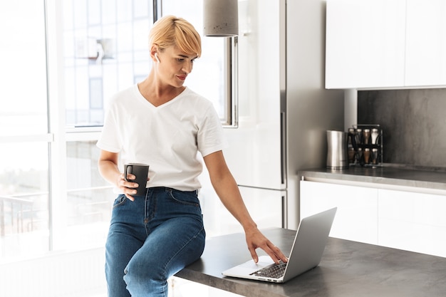 Uśmiechnięta kobieta ubrana casualy przy użyciu komputera przenośnego, siedząc w kuchni, nosząc słuchawki