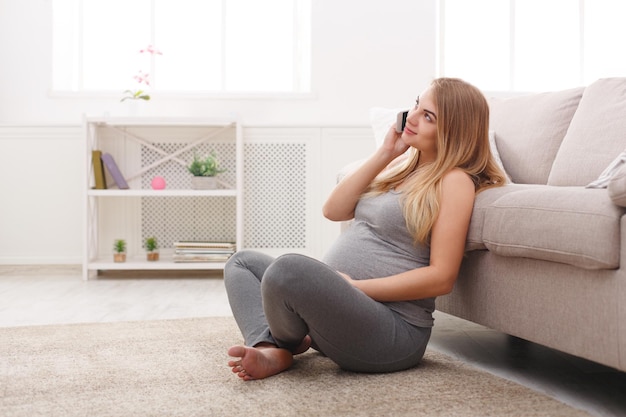 Uśmiechnięta kobieta w ciąży rozmawia na swoim smartfonie. Szczęśliwa oczekująca blondynka ma przyjemną rozmowę przez telefon i dotyka swojego brzucha, siedząc na podłodze, kopiując przestrzeń