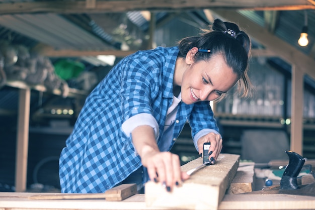 Uśmiechnięta kobieta w domowej warsztacie pomiarowej taśmy pomiarowej drewnianej deski przed piłowaniem, ciesielka