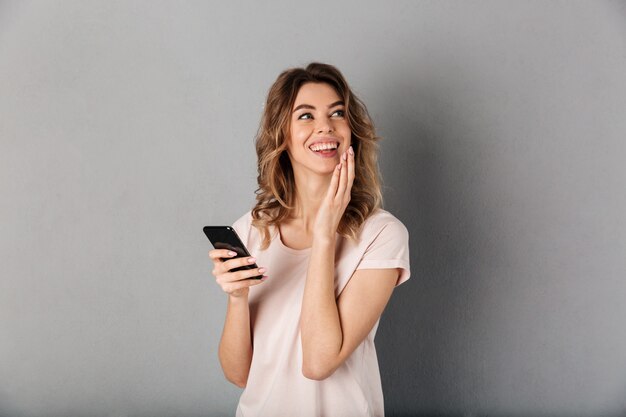 Uśmiechnięta kobieta w koszulki mienia smartphone i patrzeć daleko od nad popielatym