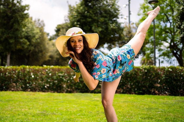 Uśmiechnięta kobieta w letniej sukience i słomkowym kapeluszu unosząca nogę podczas tańca na trawie ogrodu