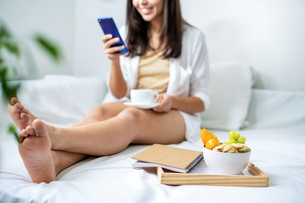 Uśmiechnięta kobieta w łóżku przy użyciu telefonu i trzymając filiżankę kawy. Śniadanie na tablicy z notatnikiem obok