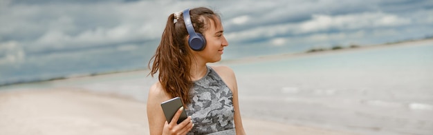 Uśmiechnięta kobieta w odzieży sportowej słuchająca muzyki na słuchawkach przy plaży na tle oceanu