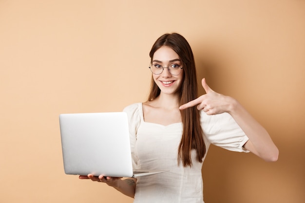Uśmiechnięta kobieta w okularach wskazując palcem na ekranie laptopa, pokazując promocję online, stojąc na beżowym tle.