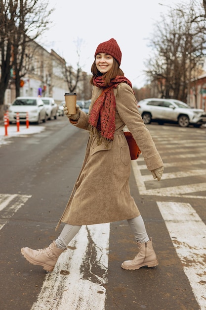 Uśmiechnięta kobieta w płaszczu spacerująca z kawą na wynos i przechodząca przez drogę jesienią