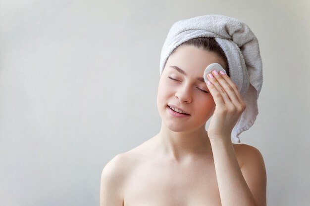 Uśmiechnięta kobieta w ręczniku na głowie z miękkim zdrowym skóry usuwać uzupełniał z bawełnianym ochraniaczem odizolowywającym na białym tle