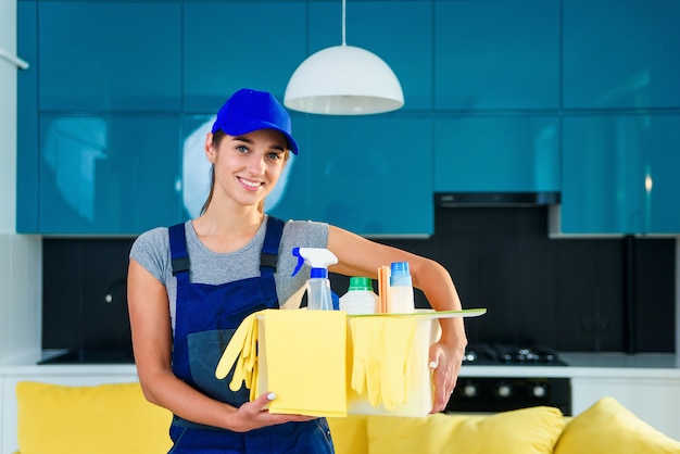 Uśmiechnięta kobieta w specjalnym mundurze do czyszczenia mieszkania trzyma pudełko z detergentami, rękawiczkami, szmatami i stoi w nowoczesnej stylowej kuchni.