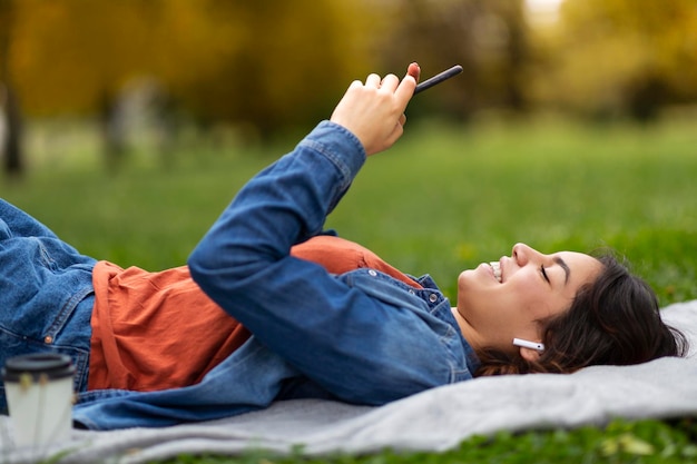 Uśmiechnięta kobieta z Bliskiego Wschodu przy użyciu smartfona, relaksując się na trawniku w parku