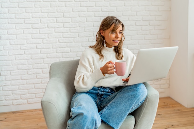 Uśmiechnięta kobieta z blond falującymi włosami siedzi na kanapie w domu, pracując na laptopie i trzymając filiżankę kawy