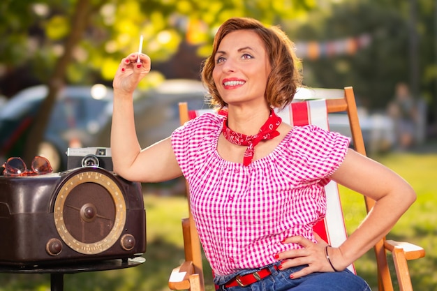 Uśmiechnięta kobieta z papierosem w ręku ubrana w styl pin-up pozuje opierając się o radio