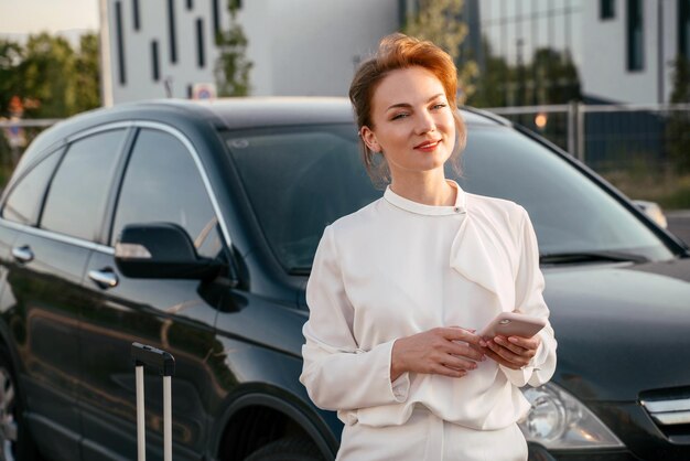 Uśmiechnięta kobieta z torbą podróżną trzymającą telefon komórkowy stojący w pobliżu samochodu. Podróż biznesowa