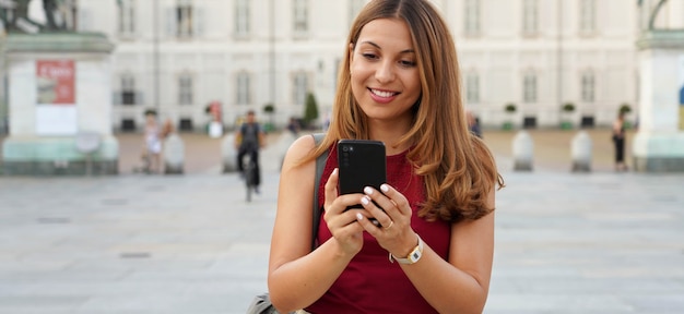 Uśmiechnięta kobieta za pomocą aplikacji telefonu komórkowego do gry online. Miasto kobieta relaks. Miejski styl życia. Upraw baner na miejsce reklamowe, tło europejskie stare miasto.