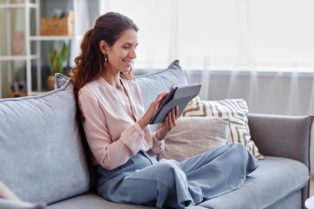 Uśmiechnięta kobieta za pomocą cyfrowego tabletu siedzi na kanapie w domu, robiąc zakupy online