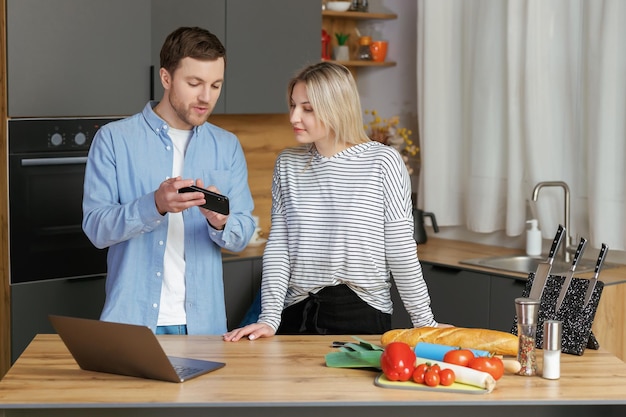 Uśmiechnięta kochająca para gotuje sałatkę razem, stojąc w kuchni w domu i używając telefonu komórkowego i laptopa