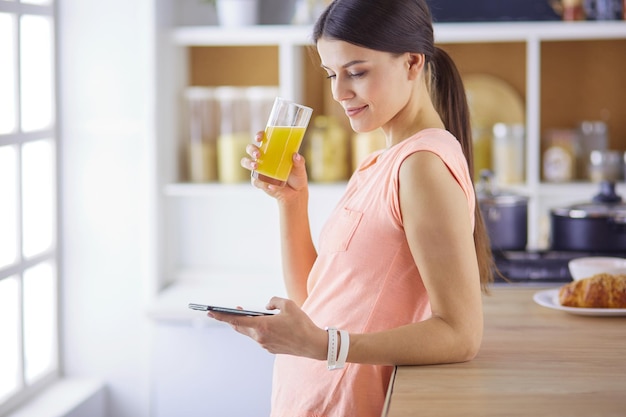 Uśmiechnięta ładna kobieta patrząca na telefon komórkowy i trzymająca szklankę soku pomarańczowego podczas śniadania w kuchni