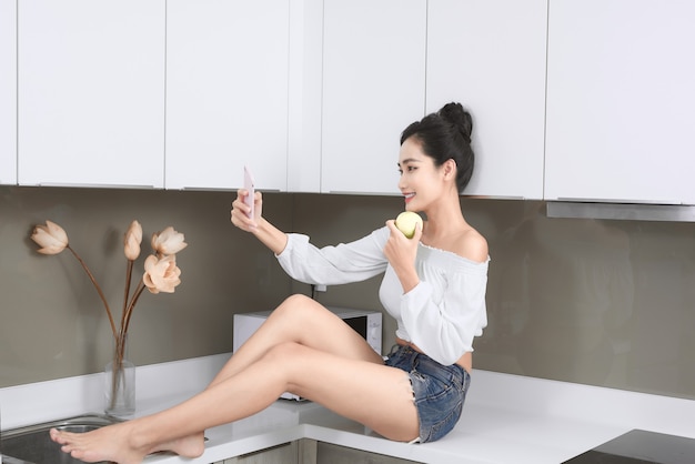 Uśmiechnięta ładna kobieta robi selfie z jabłkiem w kuchni.