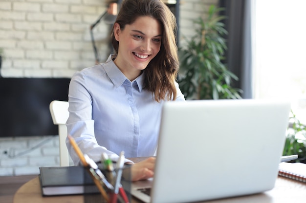 Uśmiechnięta ładna kobieta siedzi przy stole, patrząc na ekran laptopa. Szczęśliwy przedsiębiorca czytający wiadomość e-mail z dobrymi wiadomościami, rozmawiający z klientami online.