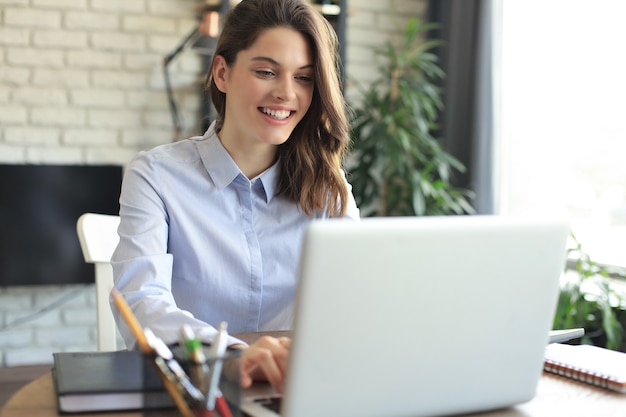 Uśmiechnięta ładna kobieta siedzi przy stole, patrząc na ekran laptopa. Szczęśliwy przedsiębiorca czytający wiadomość e-mail z dobrymi wiadomościami, rozmawiający z klientami online.