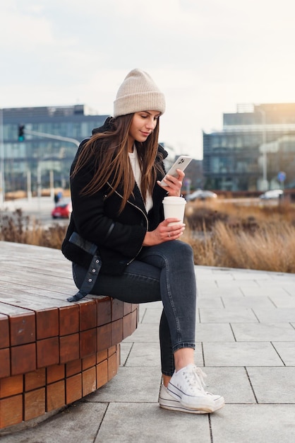 Uśmiechnięta ładna kobieta ze smartfonem i kawą, aby stać na zewnątrz i mówić nowocześnie