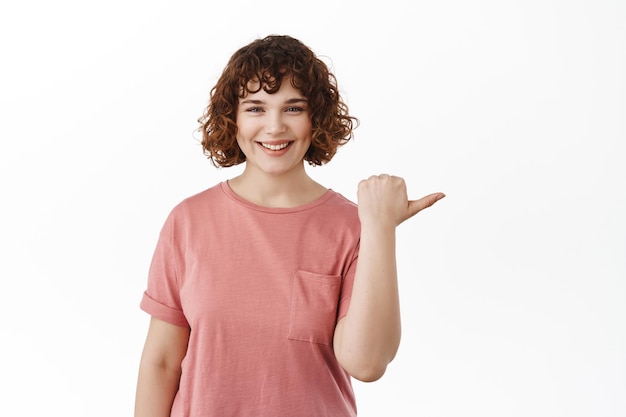 Uśmiechnięta ładna młoda dziewczyna z białym szczęśliwym uśmiechem, wskazująca kciuk w prawo, pokazująca baner reklamowy, polecająca kliknięcie ogłoszenia, stojąca w koszulce na białym tle
