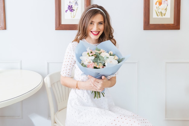 Uśmiechnięta ładna młoda kobieta w białej sukni siedzi i trzyma bukiet kwiatów w kawiarni