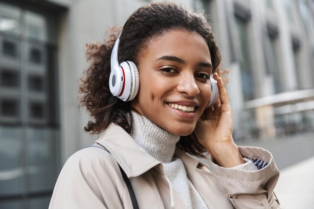 Uśmiechnięta młoda afrykańska kobieta w jesiennym płaszczu spacerująca na świeżym powietrzu, słuchająca muzyki przez słuchawki