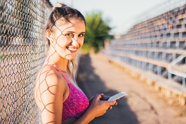 Uśmiechnięta młoda aktywna kobieta rasy kaukaskiej patrząca w kamerę i ustawiająca playlistę muzyczną na swoim telefonie