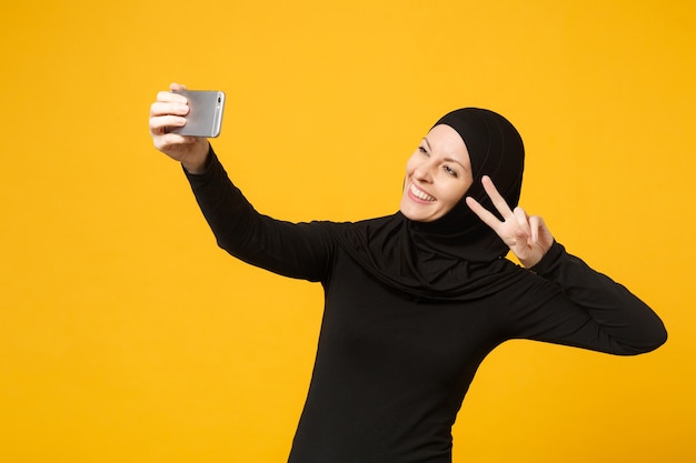 Uśmiechnięta młoda arabska muzułmańska kobieta w hidżab czarne ubrania robi selfie strzał na telefon komórkowy na białym tle na żółtą ścianę portret. Koncepcja życia religijnego ludzi. .