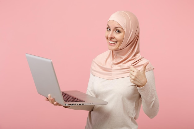 Uśmiechnięta młoda arabskiej kobiety muzułmańskiej w hidżab lekkie ubrania pozowanie na białym tle na różowym tle. Koncepcja życia religijnego ludzi. Makieta miejsca na kopię. Trzymaj i pracuj komputer typu laptop, pokazując kciuk do góry.