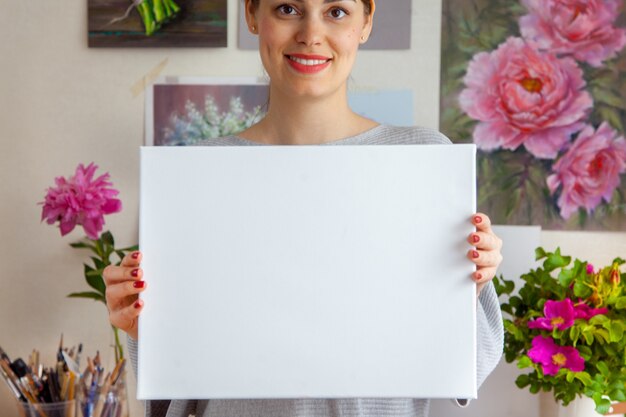 Zdjęcie uśmiechnięta młoda artystka trzyma w rękach puste białe płótno na tle przytulnego miejsca pracy z wiszącymi na ścianie obrazami i narzędziami. przestrzeń reklamowa