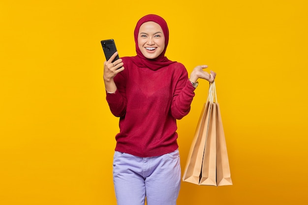 Uśmiechnięta młoda Azjatka trzymająca smartfon i trzymająca torby na zakupy na żółtym tle