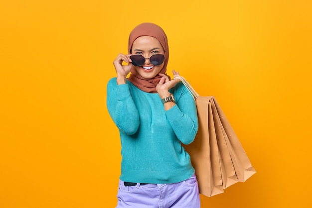 Uśmiechnięta młoda Azjatka trzymająca torby na zakupy i zdejmująca okulary na żółtym tle
