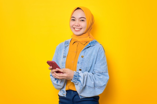 Uśmiechnięta młoda Azjatka w dżinsowej kurtce trzymająca telefon komórkowy i patrząca na kamerę odizolowaną na żółtym tle