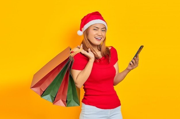 Uśmiechnięta młoda Azjatka w kapeluszu Świętego Mikołaja, korzystająca z telefonu komórkowego i trzymająca torbę na zakupy