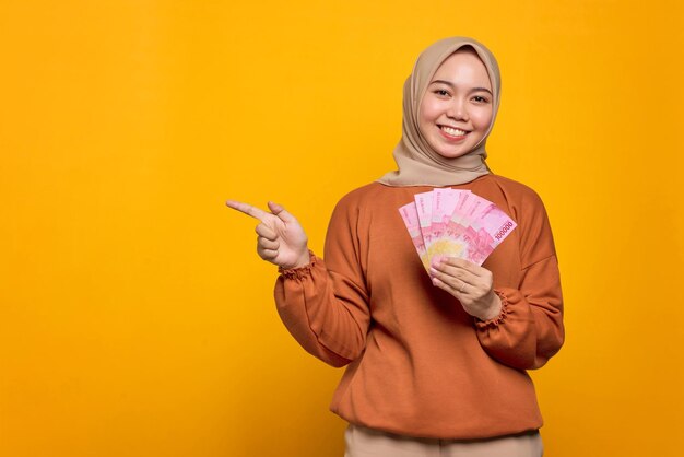 Uśmiechnięta młoda Azjatka w pomarańczowej koszuli trzymająca banknoty i wskazująca palcami na puste miejsce odizolowane na żółtym tle