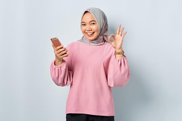 Uśmiechnięta młoda Azjatka w różowej koszuli trzymająca telefon komórkowy i pokazująca dobry znak na białym tle na białym tle