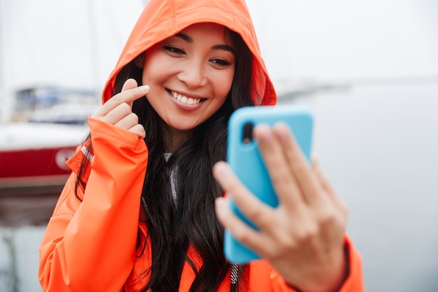 Uśmiechnięta młoda azjatycka kobieta ubrana w płaszcz przeciwdeszczowy spędzająca czas na świeżym powietrzu, spacerująca po wybrzeżu, robiąca selfie