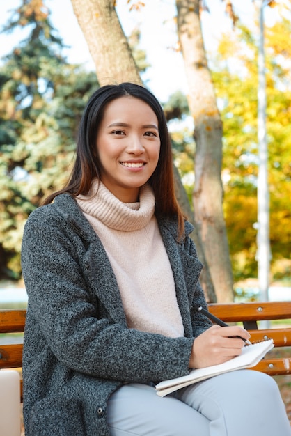 Uśmiechnięta Młoda Azjatycka Kobieta Ubrana W Płaszcz Siedzi Na ławce W Parku, Robiąc Notatki W Notatniku