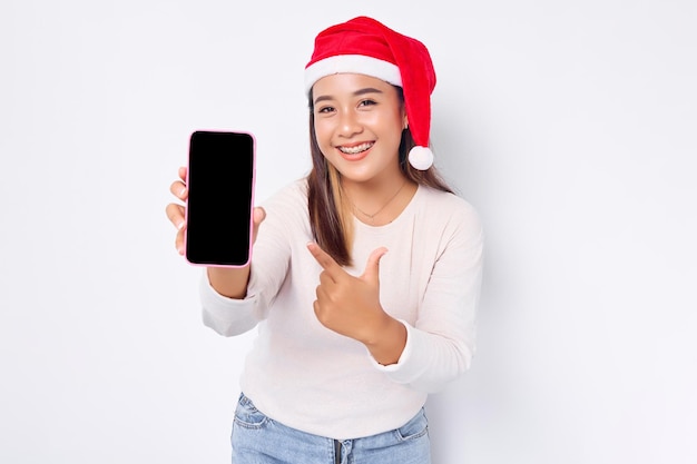 Uśmiechnięta młoda azjatycka kobieta w świątecznym kapeluszu pokazująca pustego ekranu telefon komórkowy polecająca aplikację mobilną izolowaną na białym tle