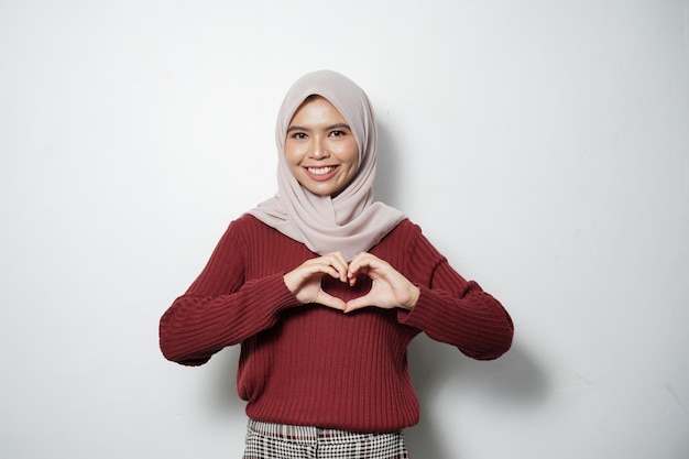 Uśmiechnięta młoda azjatycka muzułmanka pokazująca gest serca robi znak miłości palcami odizolowanymi na białym tle